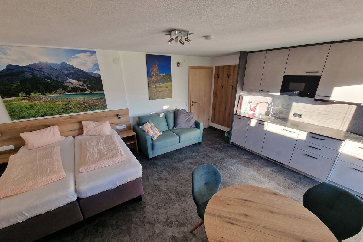 Ein modernes Zimmer mit Bett, einer Couch und einem Esstisch. Es gibt eine Küchenzeile mit Schränken und einer Mikrowelle.