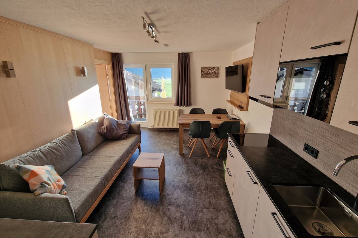 Ein kompakter Wohnraum mit Sofa, kleinem Tisch, Essbereich mit Stühlen, einer Küche mit Spüle und Herd sowie einem an der Wand montierten Fernseher.