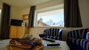 Un comedor con una mesa de madera, una tabla de cortar con comida, una copa de vino y un control remoto. Hay un televisor sobre un mueble de madera y una ventana con vistas a las montañas.