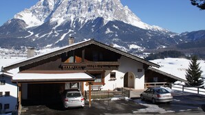 A mountain lodge with a garage and two cars parked in front, set against a backdrop of snow-covered mountains. - Ferienhaus Mott (Lermoos)