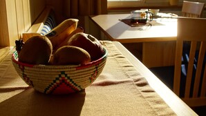 Una mesa de madera con una cesta tejida que contiene pan, una ventana con vista a la montaña y una silla de madera.