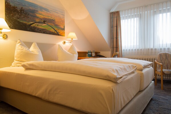A neatly made bed with white linens, a painting of a landscape on the wall, and a window with sheer curtains. - Hotel-Restaurant Bastenhaus (Dannenfels)