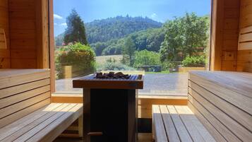 A wooden cabin with a table and benches, a view of a forested hillside, and a clear sky.