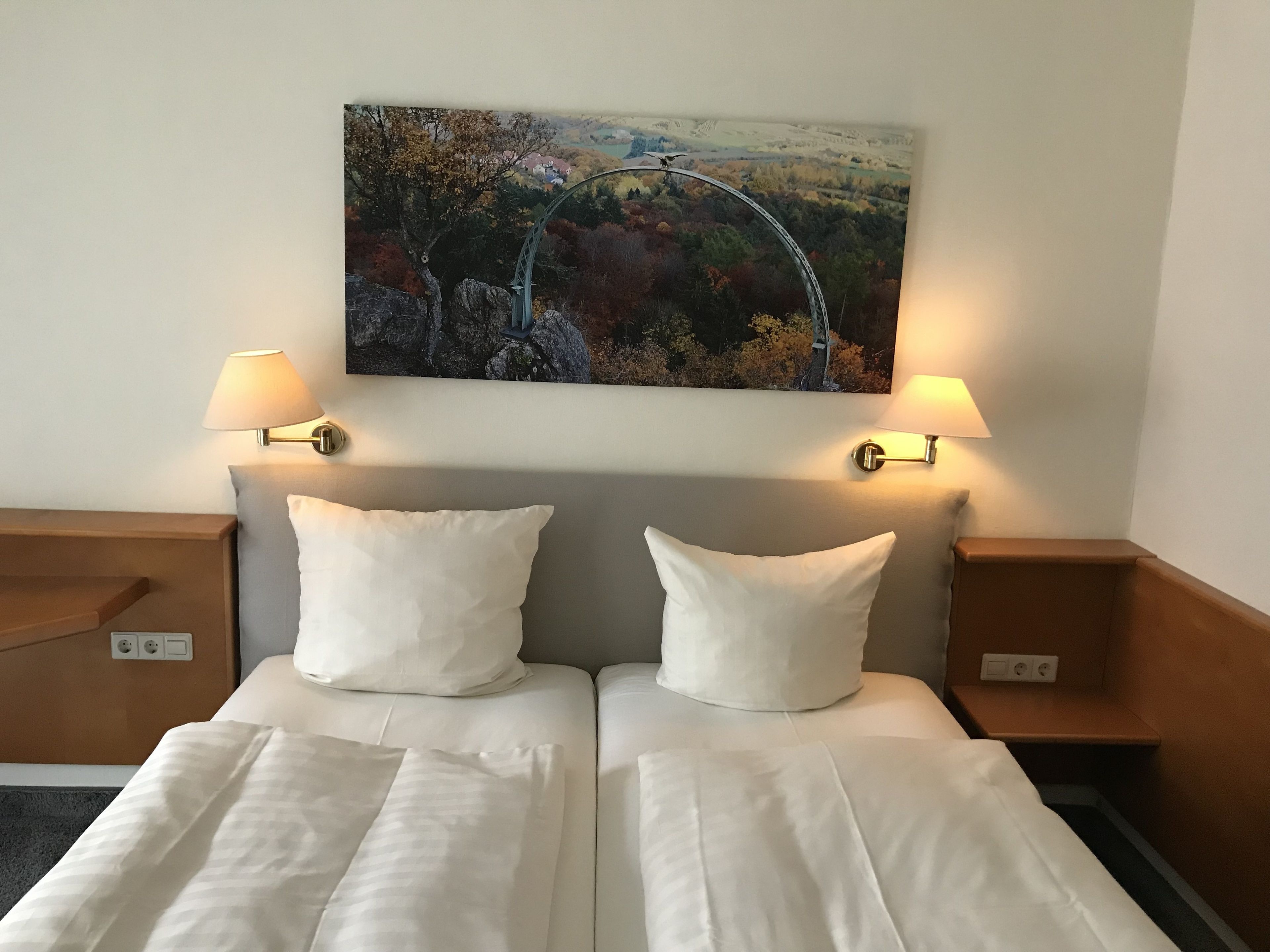 A hotel room with two beds, each with a white pillow and a white bedspread. There are two bedside tables with lamps, and a painting of a landscape hangs above the beds.