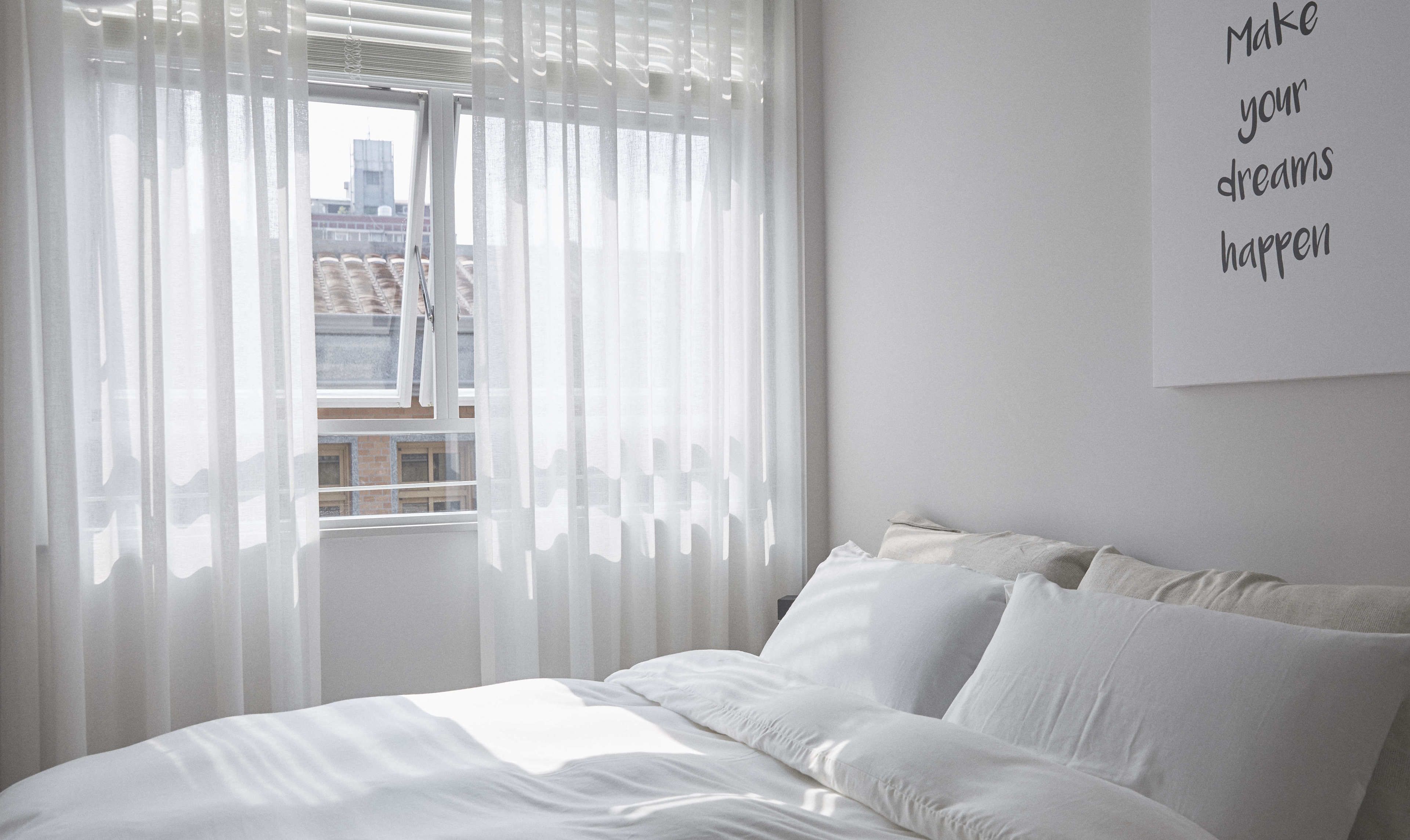 A bedroom with a bed, white curtains, and a city view through the window.