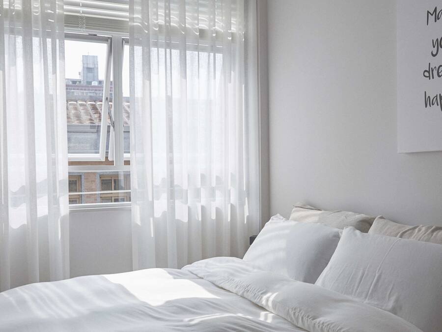 A bedroom with a bed, white curtains, and a city view through the window.