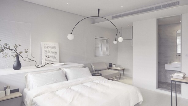 Een moderne slaapkamer met een groot bed, een bureau en een stoel. Er is een raam met jaloezieën, een vaas met takken en een wandplank.