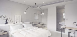A modern bedroom with a large bed, a desk, and a chair. There is a window with blinds, a vase with branches, and a wall-mounted shelf.