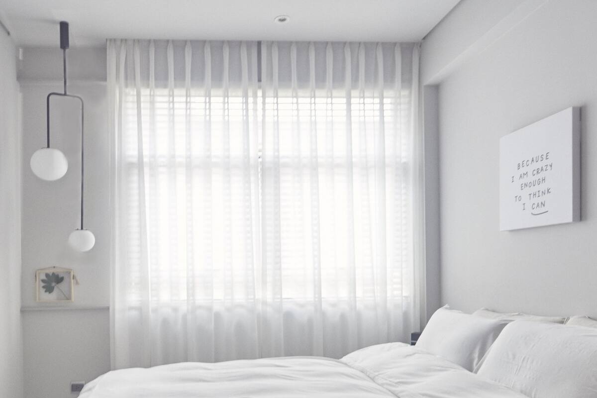 A minimalist bedroom with a bed, a television, a wall-mounted clock, and a window with sheer curtains.