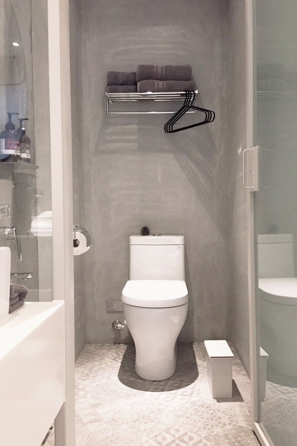 Une salle de bain avec des toilettes, une étagère au-dessus des toilettes, un petit meuble blanc et un sol à motifs.