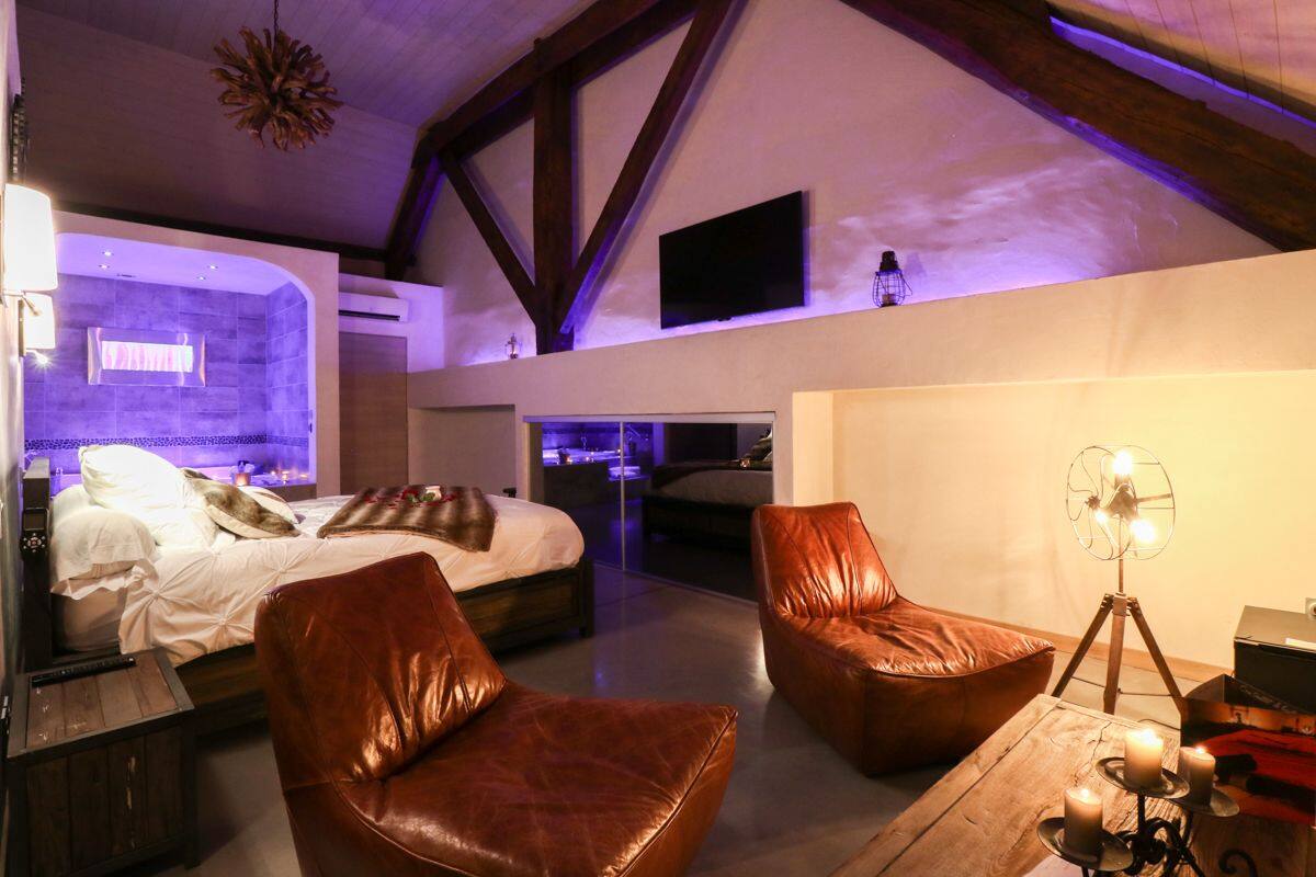 A modern bedroom with a large bed, a seating area with leather chairs, a wooden coffee table, and a wall-mounted TV.