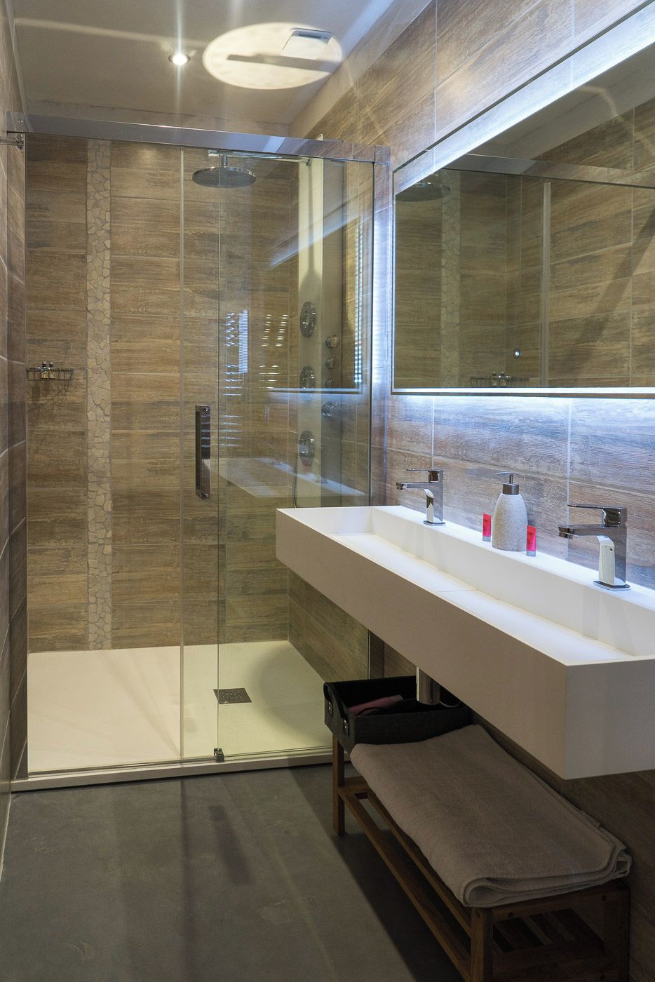 Un baño moderno con ducha envuelta en vidrio, un espejo grande y un tocador blanco con banco.