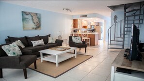 Relax in this stylish living space with a sofa, coffee table, and armchair, plus a kitchen area and staircase leading to an upper level.