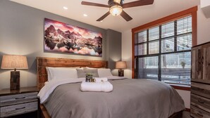 A bedroom with a large bed, wooden headboard, two bedside tables with lamps, a ceiling fan, a large window with blinds, and a painting on the wall. - Modern Condo in Heart of Village, 1st floor, Steps to Gondola & all Amenities   (Mammoth Lakes)