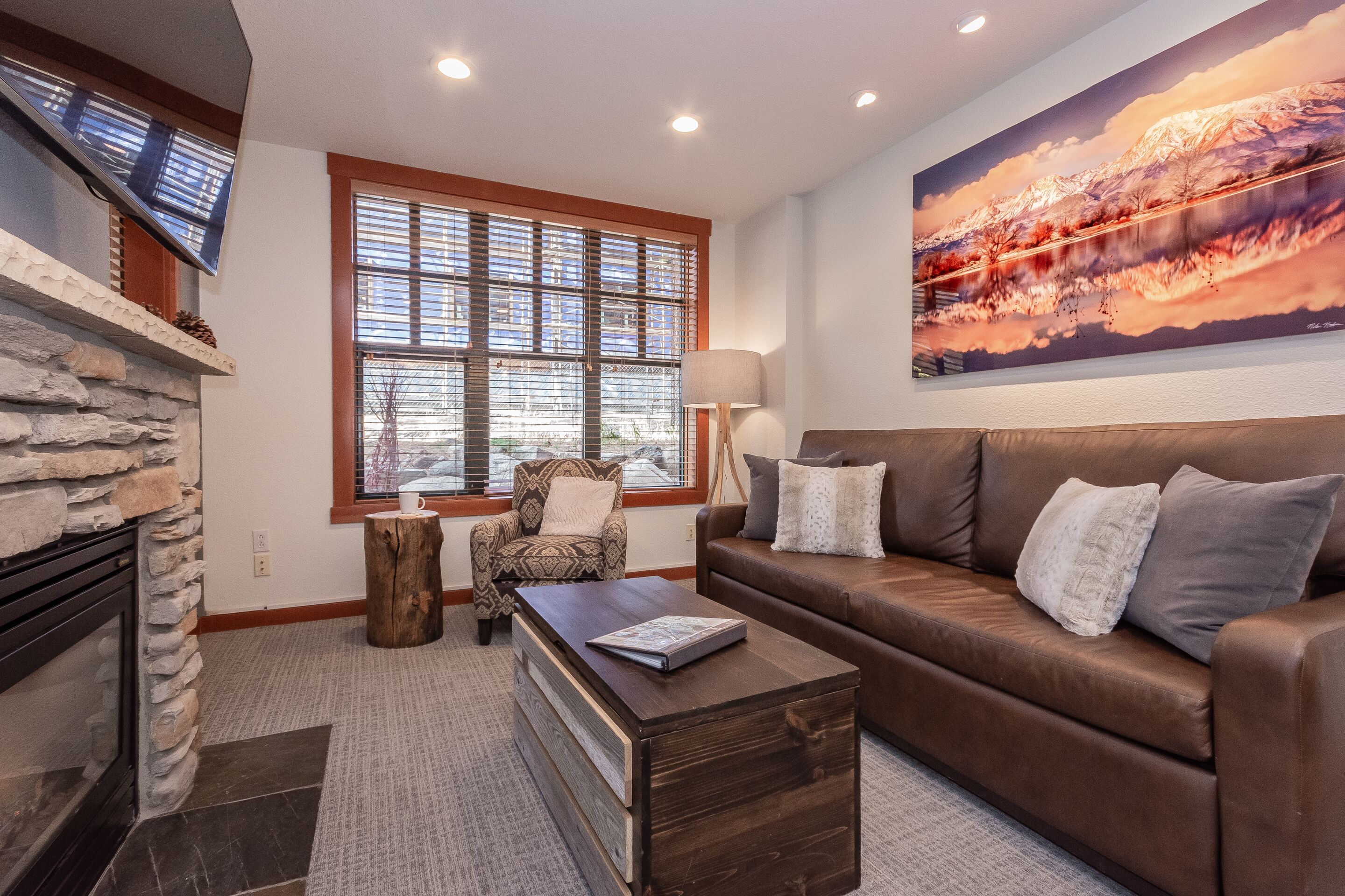 A living room with a stone fireplace, a brown leather sofa, a wooden coffee table, and a large landscape painting on the wall.