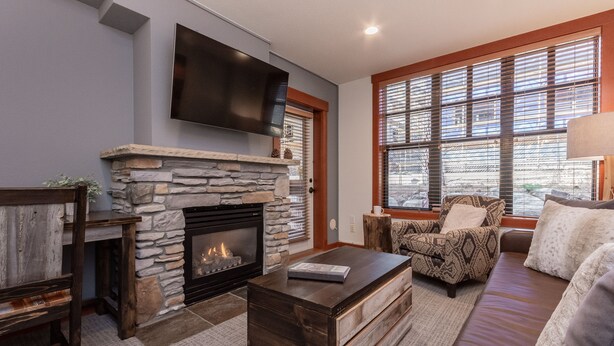 A living room with a stone fireplace, a flat-screen TV mounted above it, a sofa, a patterned armchair, and a coffee table. - Modern Condo in Heart of Village, 1st floor, Steps to Gondola & all Amenities   (Mammoth Lakes)
