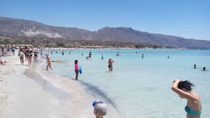 Une plage bondée, avec une eau turquoise et limpide, et des montagnes en arrière-plan.