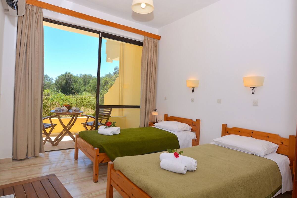 A hotel room with two beds, a balcony with a table and chairs, and a view of greenery.