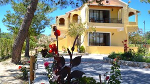 A two-story building with a yellow facade, multiple balconies, and a garden with various plants and flowers. - Alexandra Apartments (Corfu)