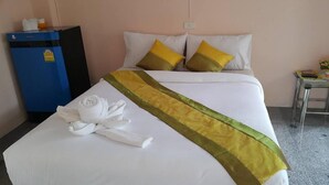 Una cama bien hecha con sábanas blancas y una franja decorativa verde y amarilla, un minibár azul y una pequeña mesa con un control remoto y un florero con flores.