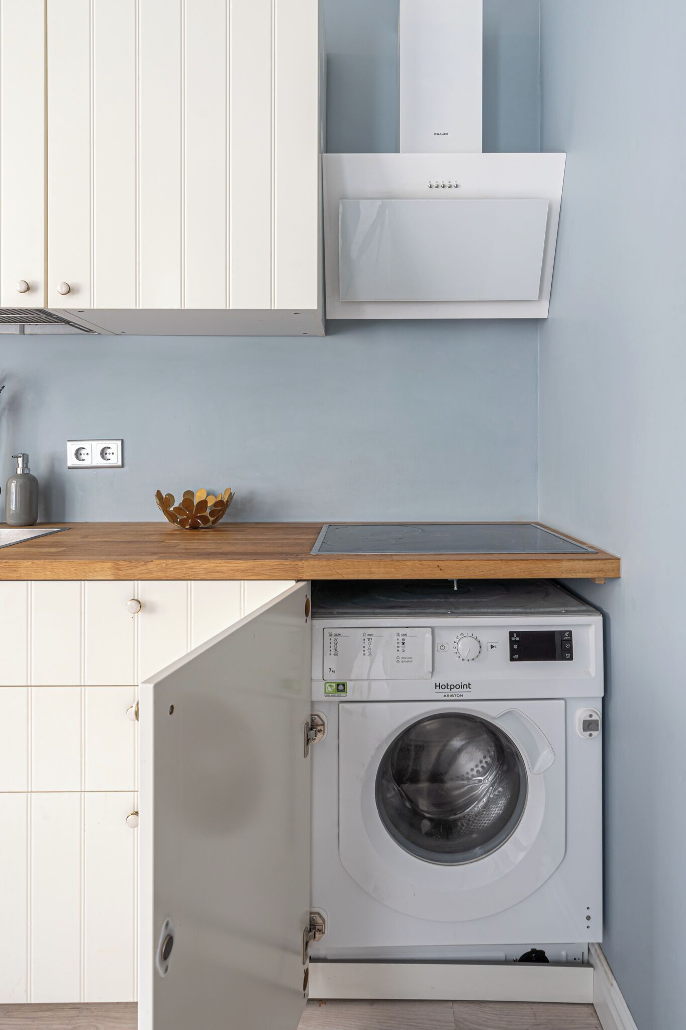 A compact kitchen with a built-in washing machine, a wooden countertop, and white cabinetry.