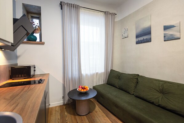 A compact living space with a kitchenette, a green sofa, a round table with a fruit bowl, a window with curtains, and wall art.