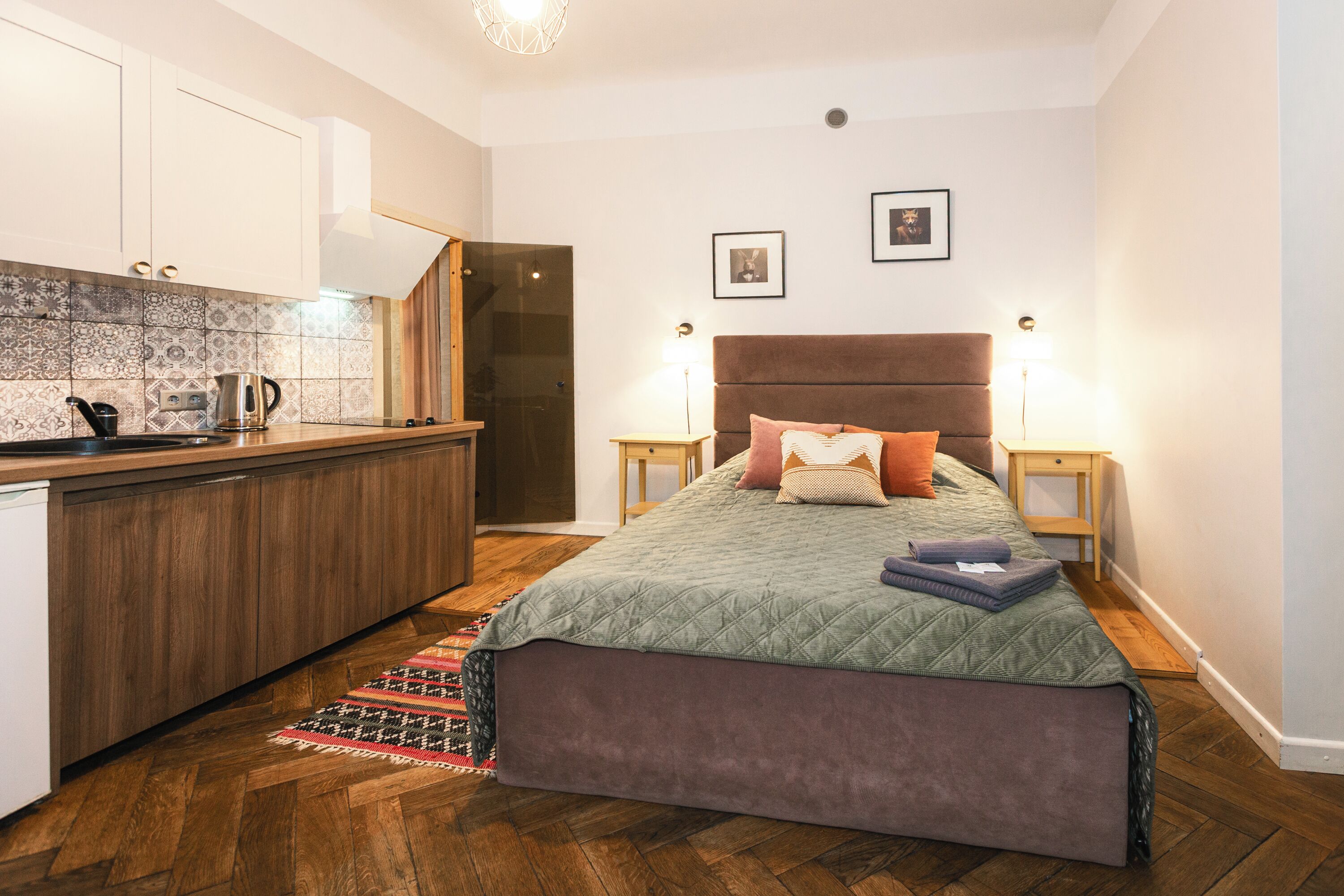 A bedroom with a bed, bedside tables, a nightstand, a wooden floor, a kitchen area with cabinets and a countertop, and a patterned backsplash.