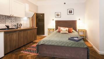 A bedroom with a bed, bedside tables, a nightstand, a wooden floor, a kitchen area with cabinets and a countertop, and a patterned backsplash.