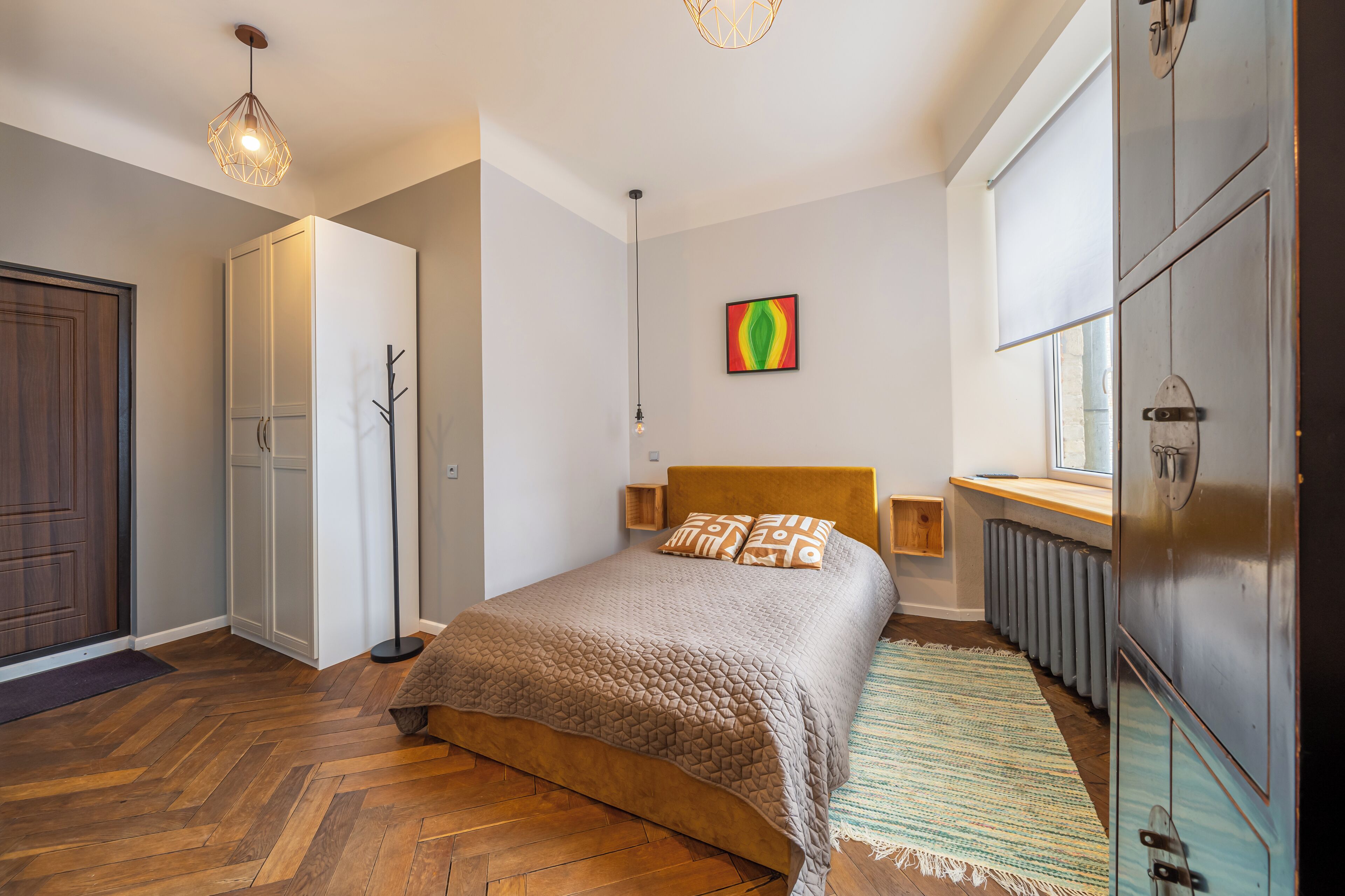 Un dormitorio con piso de madera, una cama con colcha marrón, un radiador y un ventanal con persianas.
