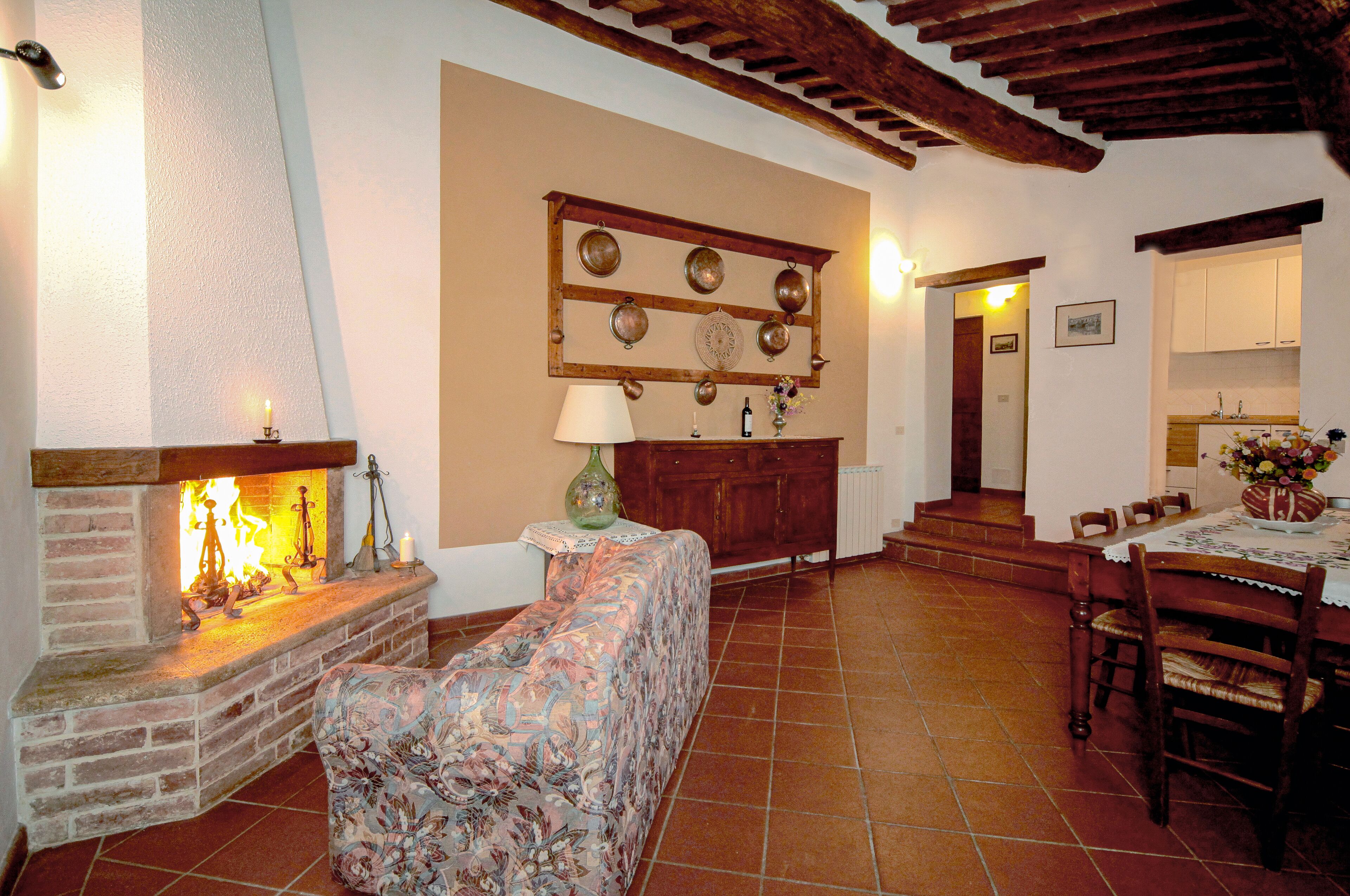 A cozy living room with a fireplace, a sofa, a dining table with chairs, and a kitchen area in the background.