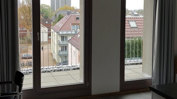 Une pièce dotée de deux grandes fenêtres, d’un fauteuil et offrant une vue sur des immeubles et une rue.