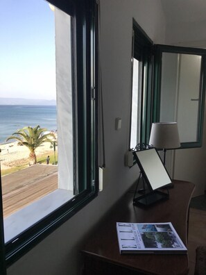 A room with a large window offering a view of the beach and ocean. There is a desk with a lamp and a magazine on it.