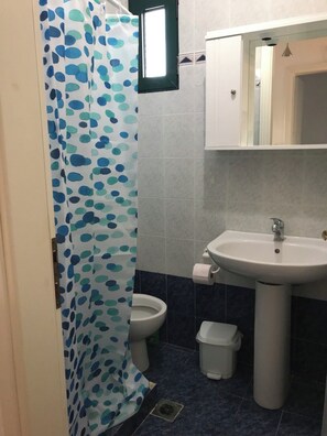 A bathroom with a shower curtain, a toilet, a sink, and a trash bin. - Stunning sea-front maisonette (Kassandra)