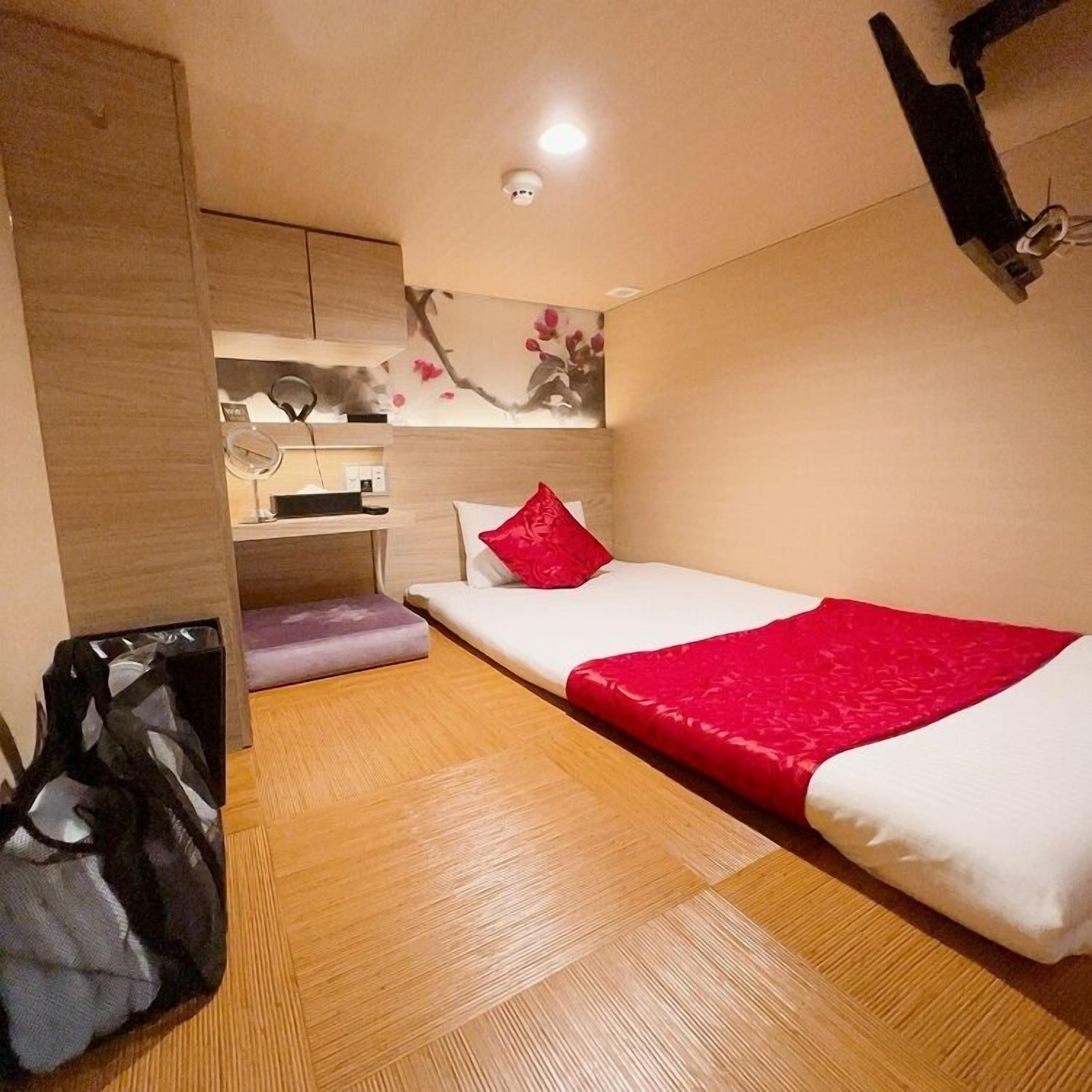 A compact hotel room with a single bed, a small desk, a wardrobe, and a ceiling-mounted light fixture.