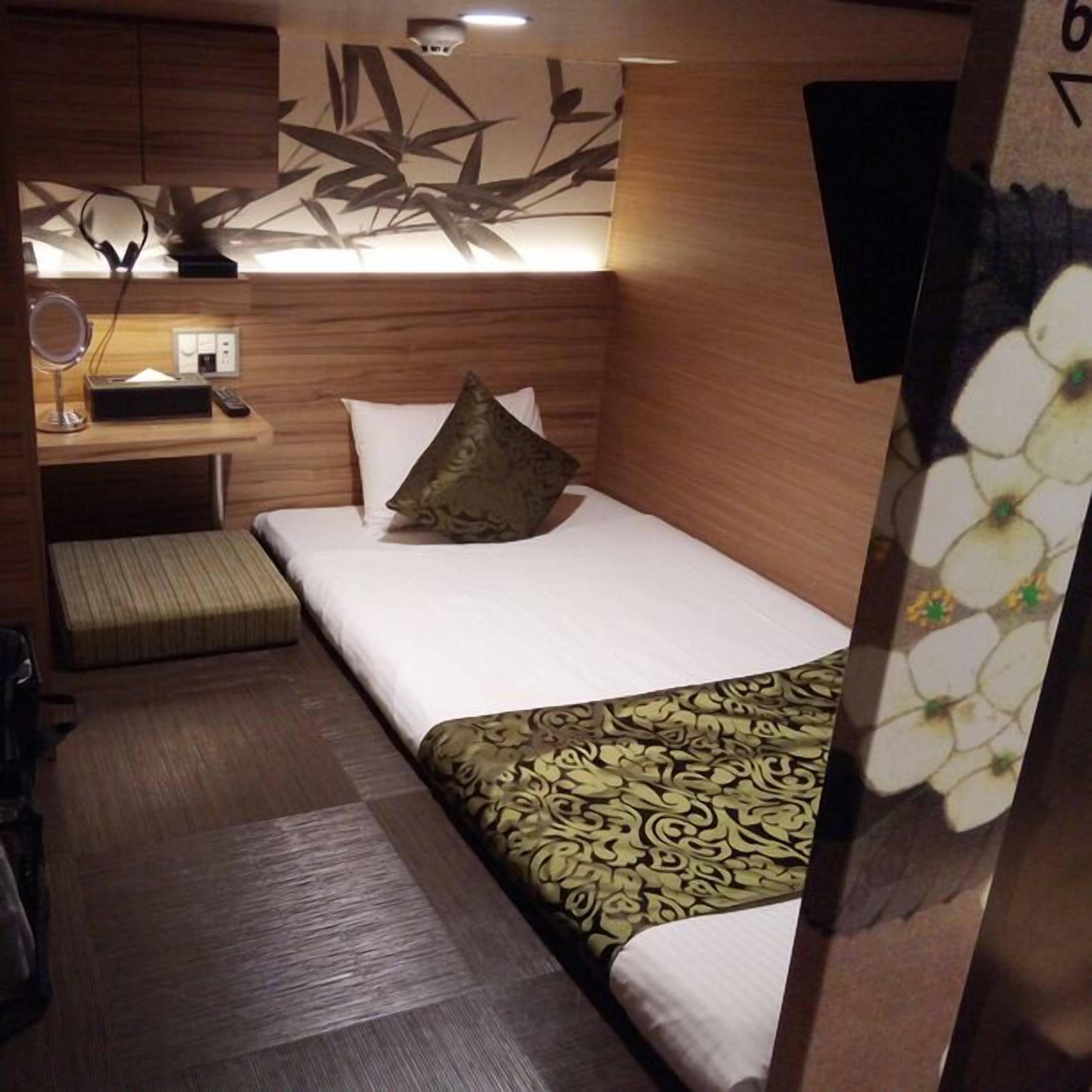 A compact hotel room with a bed, a small desk with a lamp, a mirror, and a wall-mounted light fixture.