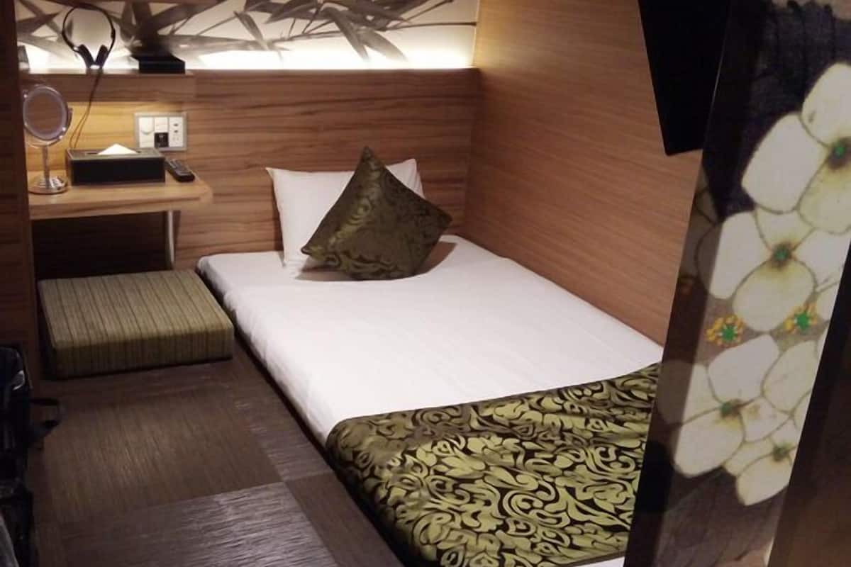 A compact hotel room with a bed, a small desk with a lamp, a mirror, and a wall-mounted light fixture.