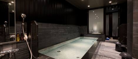 A modern spa with a large rectangular hot tub, dark wooden paneling, and a glass-enclosed shower area.