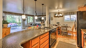 A spacious kitchen with granite countertops, wooden cabinets, and a large island. There are pendant lights, a refrigerator, and a dining area with chairs and a table. - Hot Tub + Fireplace: Peaceful Anderson Island Home (Anderson Island)