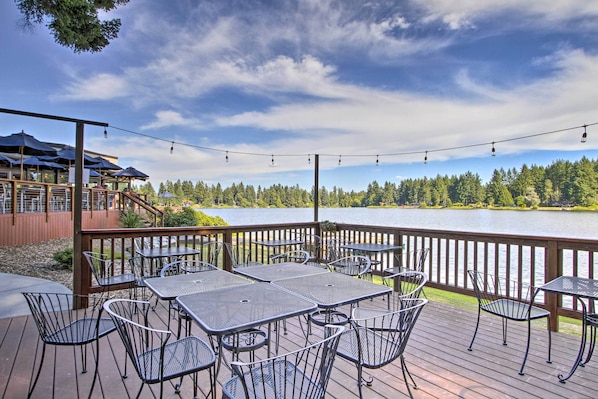 A deck with tables and chairs overlooking a lake, surrounded by trees and a wooden deck railing.