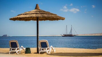 A beach with two white lounge chairs and a thatched umbrella. A sailboat is visible in the distance on the water.