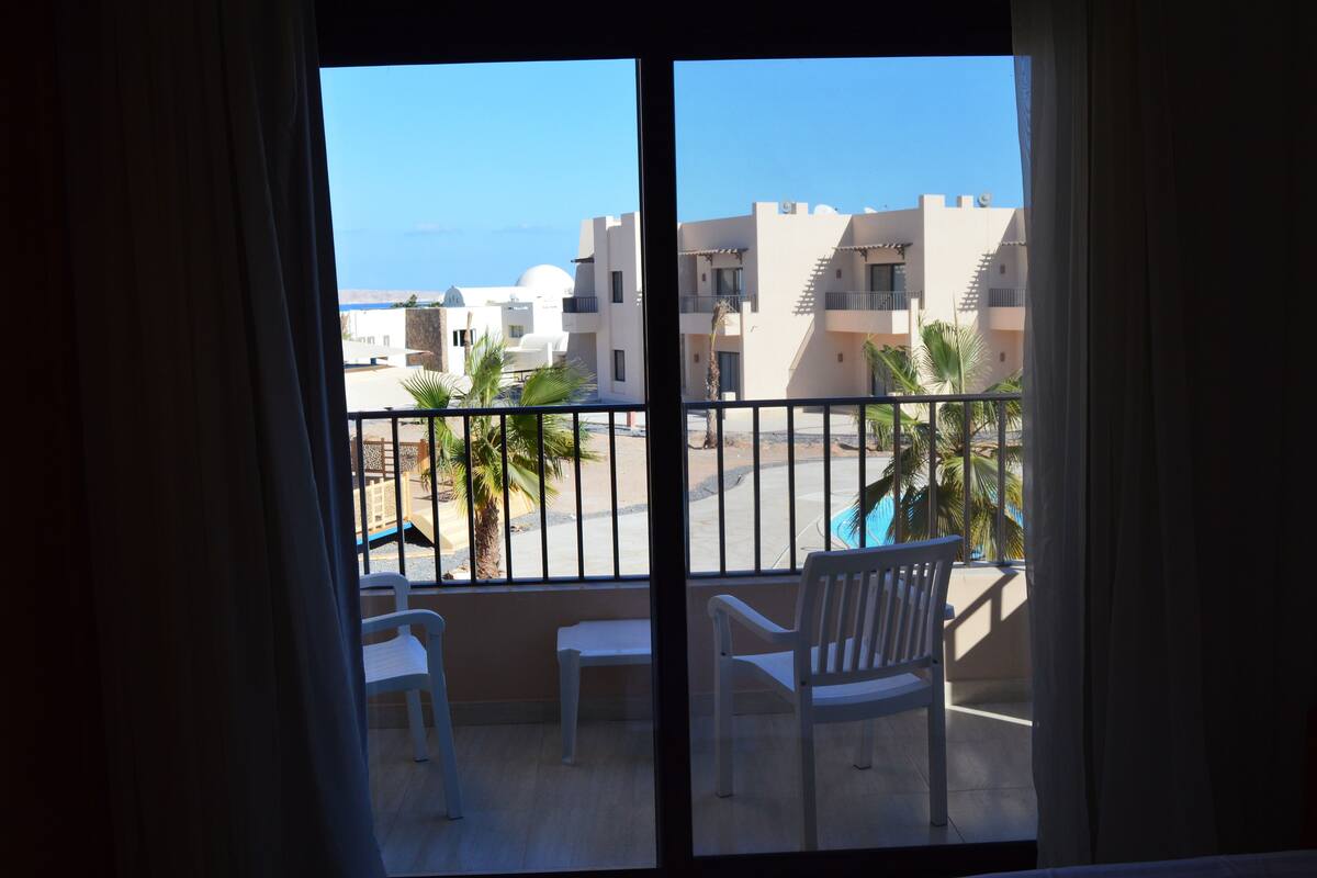 A balcony with white chairs and a small table, overlooking a pool and buildings with a clear view of the sea.