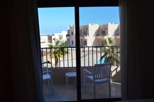 A balcony with white chairs and a small table, overlooking a pool and buildings with a clear view of the sea.