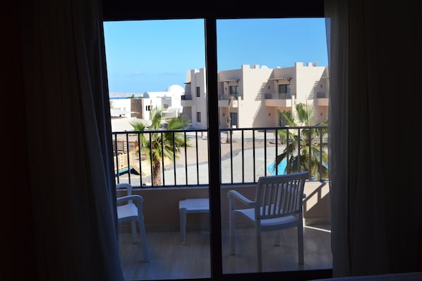 Un balcón con sillas blancas y una pequeña mesa, con vistas a una piscina y edificios, y una vista despejada del mar.