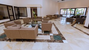 Un vestíbulo amplio con varias zonas de estar, una mesa de cristal central y grandes ventanales que ofrecen vistas a palmeras.