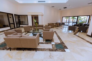 A spacious lobby with multiple seating areas, a central glass table, and large windows offering views of palm trees.
