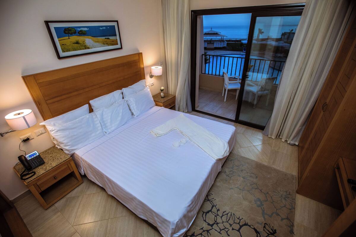 A hotel room with a large bed, a nightstand with a phone, a painting on the wall, and a balcony with chairs.