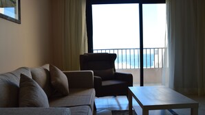 Una sala de estar con sofá, sillón y mesa de centro. Se ve el mar a través de la puerta del balcón.