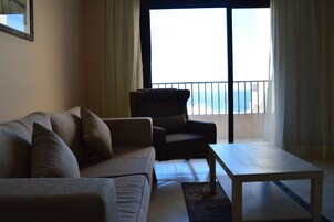 A living room with a sofa, armchair, and coffee table. There is a view of the sea through a balcony door.
