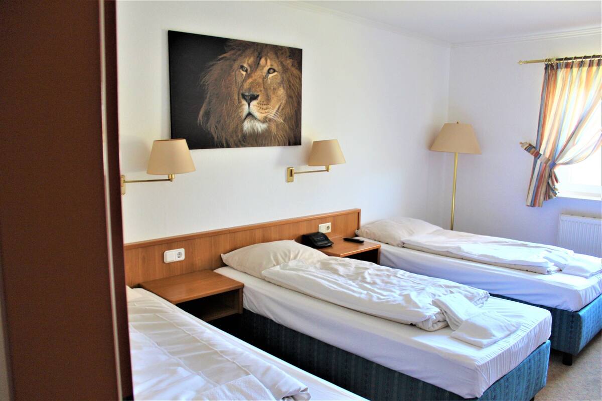 A hotel room with two single beds, a wooden headboard, a wall-mounted lamp, a bedside table with a phone, and a painting of a lion.