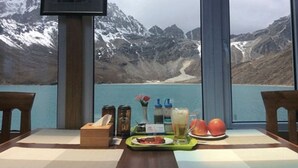 Una mesa con comida, una botella y un vaso, frente a un paisaje montañoso que se ve a través de una ventana.
